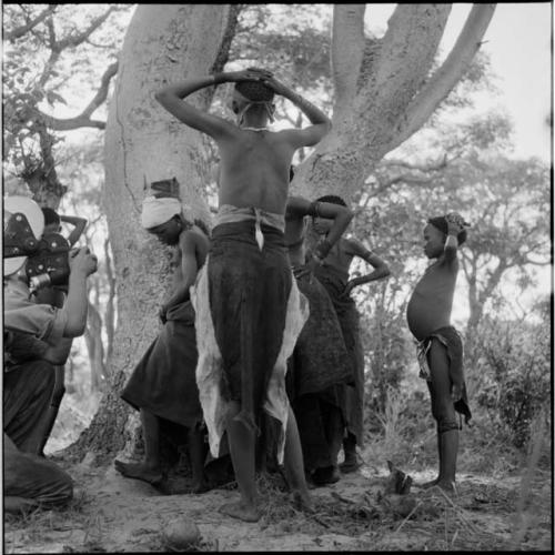 Group of women and girls playing Tcxai Djxani (a dancing game) at a tree, with N!ai dancing in a hole made by their feet, two girls with their hands on the heads, John Marshall filming them