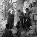 Group of women and girls playing Tcxai Djxani (a dancing game) at a tree, with N!ai and another girl clapping, John Marshall filming them