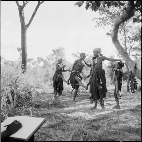 Group of women and girls playing Tcxai Djxani (a dancing game), dancing in a line, with N!ai at the end of the line, clapping