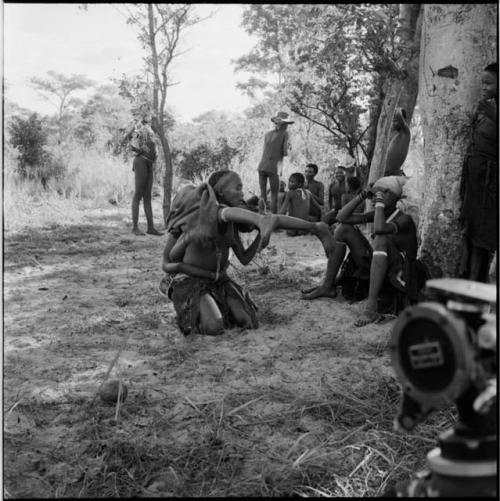 Group of women and girls playing Tcxai Djxani (a dancing game) at a tree, with one girl putting another girl on her shoulders, John Marshall filming them