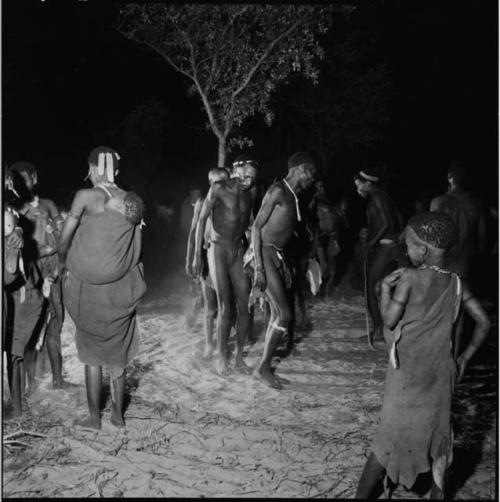 Men dancing in a line in a night dance, with women carrying babies on their backs standing near them