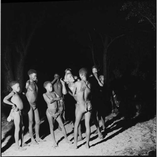 Men dancing in a line through a group of boys standing, in a night dance