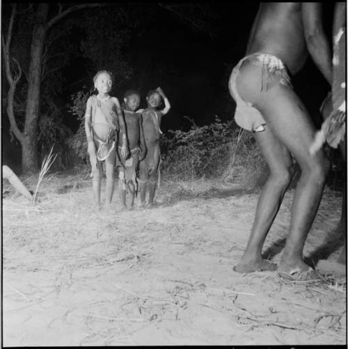 Three children standing, watching a group of dancers in a night dance