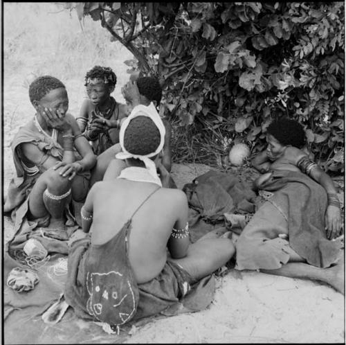 Woman wearing a decorated bag across her back, sitting with a group of women next to a shelter skerm