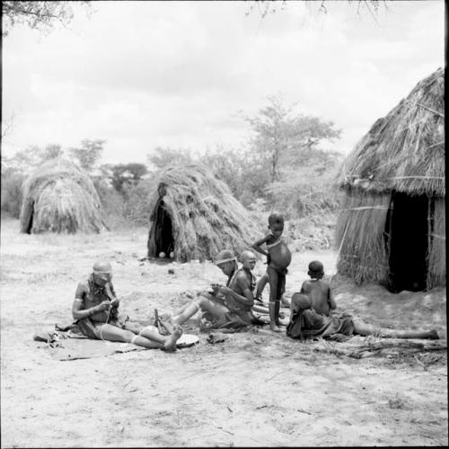 Group of women and children sitting, including a boy playing a //guashi and a woman stringing ostrich eggshell beads