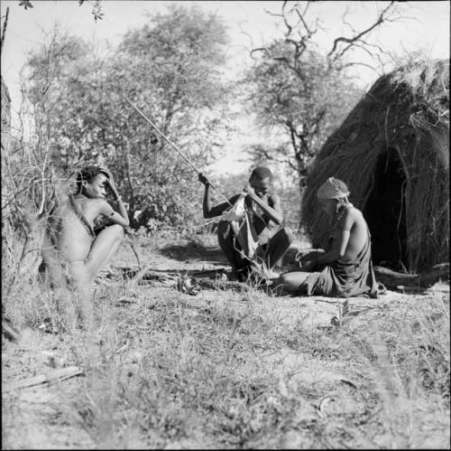 Man putting a spear in his bag, sitting with his wife in front of a skerm, with a man squatting near them, dog sitting next to them