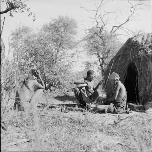 Man putting a spear behind him, sitting with his wife in front of a skerm, with a man squatting near them, dog sitting next to them