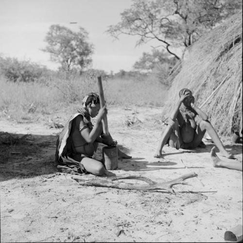 Woman pounding something in a mortar and pestle, sitting with another woman near a skerm