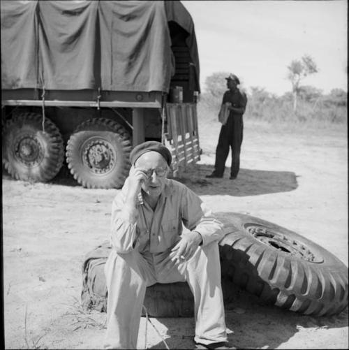 Charles Koch sitting on a tire covered with canvas, with another expedition member standing next to a truck in the background