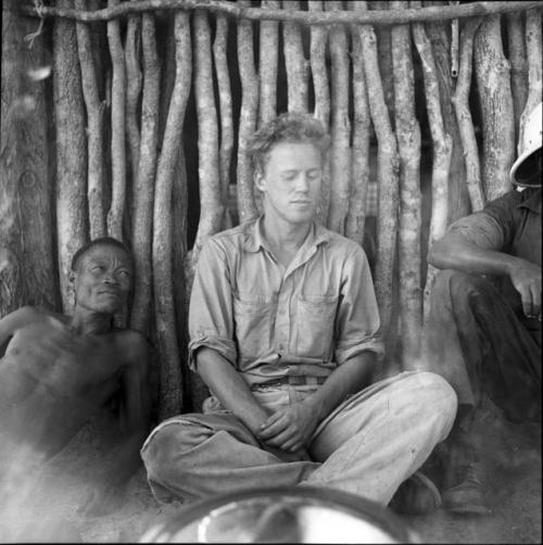 /Ti!kay and another man sitting next to John Marshall in front of one of the walls of the expedition hut