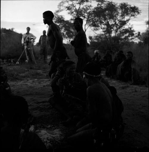 /Ti!kay and another man dance by groups of people sitting, with John Marshall and Laurence Marshall in the background