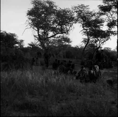 /Ti!kay and two other men dancing next to several groups of people sitting at a daytime dance, distant view