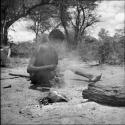 Man sitting next to a fire, cracking mangetti nuts with a rock