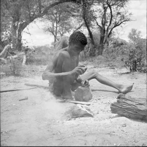 Man sitting next to a fire, cracking mangetti nuts with a rock