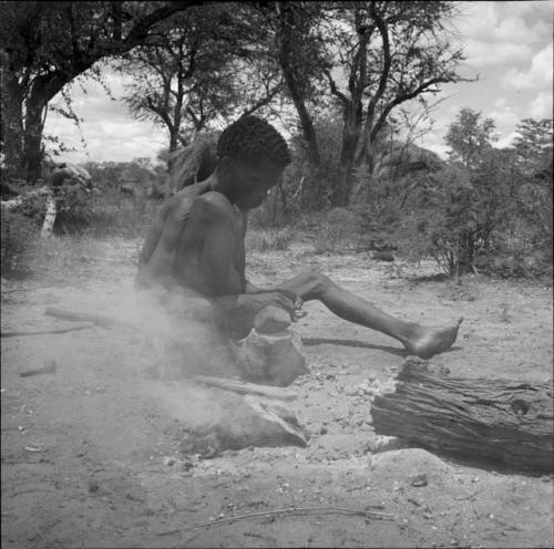 Man sitting next to a fire, cracking mangetti nuts with a rock