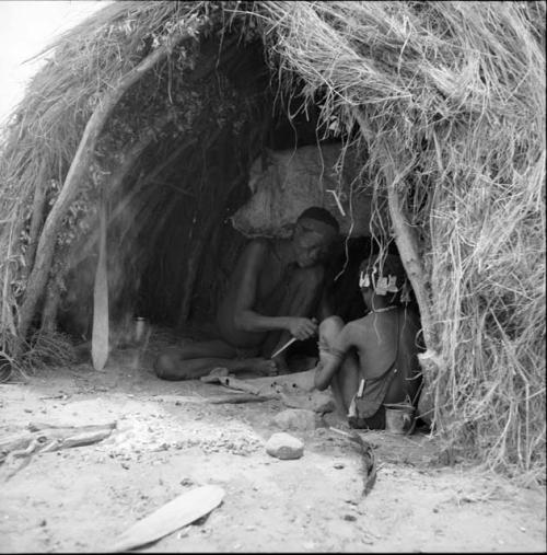 Man cutting strips of a dried hide, sitting inside a skerm with a girl, fire paddles on the ground next to them