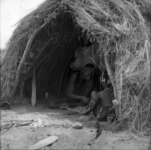 Man cutting strips of a dried hide, sitting inside a skerm with a girl, fire paddles on the ground next to them