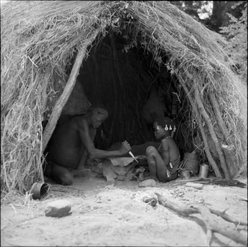 Man cutting strips of a dried hide, sitting inside a skerm with a girl, dance rattles hanging inside the skerm