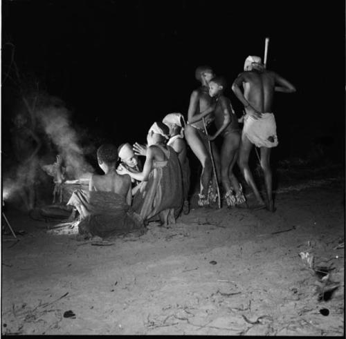 Boys dancing in a night dance near a group of women sitting and clapping, with smoke from two fires behind them