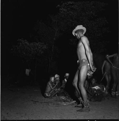 Man wearing dance rattles and a Western hat dancing in a night dance, with a group of women and a boy siting and clapping behind him