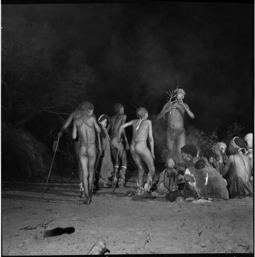 Boys dancing in a night dance, one holding a tree branch, near a group of women with babies sitting and clapping