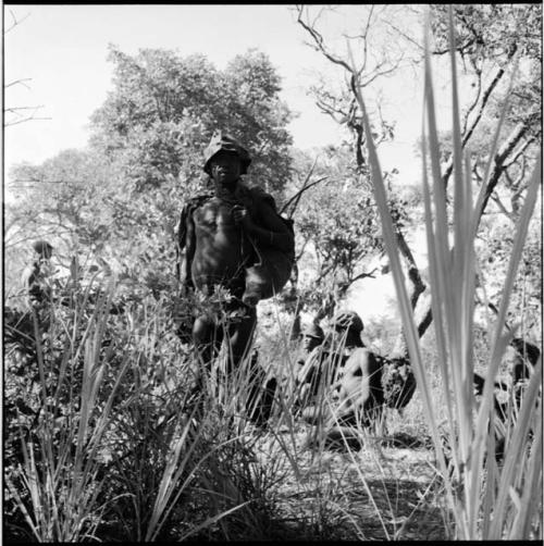 Man carrying hunting equipment standing, with other hunters sitting next to bushes behind him