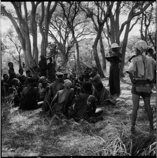 Group of women sitting next to a tree, including "Old /Naoka" and a woman wearing a printed dress, with John Marshall filming them, expedition members standing near them