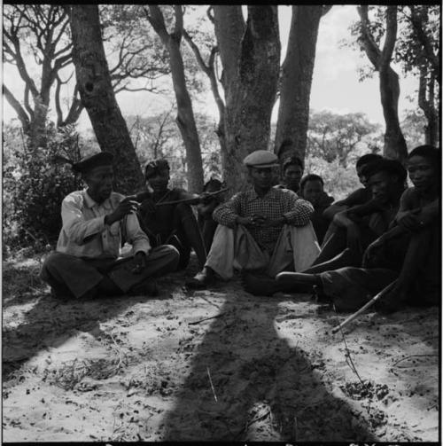 "Short /Qui" sitting with a group of people, including a man playing a musical bow