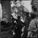 Group of men, including /Gunda, standing next to a tree, talking with John Marshall holding his film camera