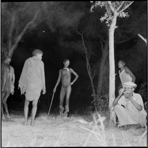 Group of people dancing in a night dance, including a man wearing dance rattles and a boy wearing a kaross