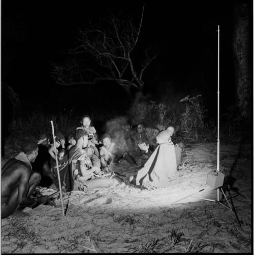 Man chopping with an axe, squatting next to a group of women sitting around a fire, clapping and singing