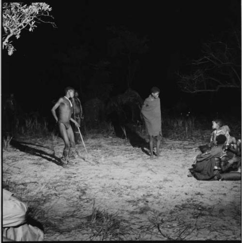 Man wearing dance rattles and carrying a stick, dancing near a group of women and children sitting and clapping in a night dance, with a boy wrapped in a blanket standing near them