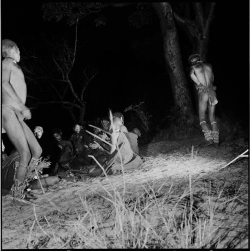 Two men dancing by a group of women sitting, clapping and singing for a night dance