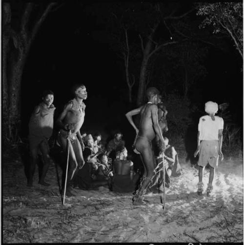 Five men dancing by a group of women sitting, clapping and singing for a night dance, with one man wearing Western clothes