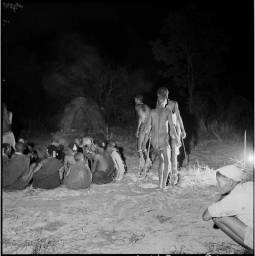 Men dancing by a group of women sitting, clapping and singing for a night dance, with skerms in the background