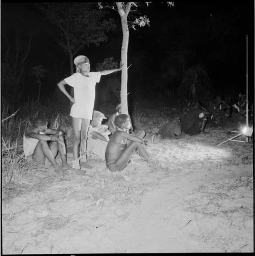 Boys sitting and standing next to a tree, watching a night dance