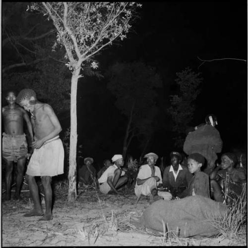 Two men standing, one smoking a pipe, with a group of people sitting near them