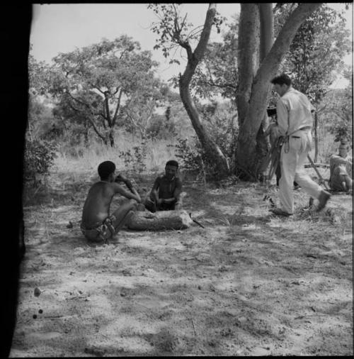 !Naishi sitting and eating, with //Ao sitting next to him chopping at a log, making a //guashi, an expedition member walking toward them