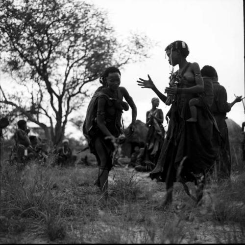 Two women with babies on their backs dancing and clapping, playing tamah n!o’an (a ball game) / !’hu kuitzi (veldkos game)