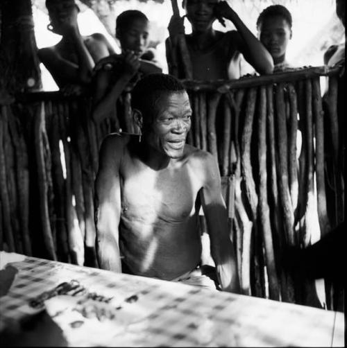 /Ti!kay sitting at a table inside the expedition hut, with beetle specimens on the table in front of him, people standing outside behind him