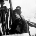 ≠Toma wearing a coat, sitting at a table in the expedition hut, with two people standing behind him