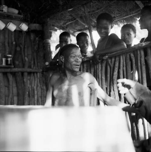 /Ti!kay sitting at a table in the expedition hut, talking, with a group of men and boys, including /Gaishay, standing outside behind him, listening