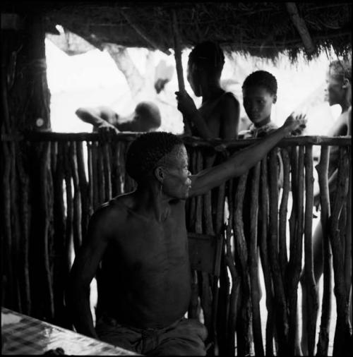 /Ti!kay sitting at a table in the expedition hut, pointing at the fence behind him, looking at a group of men and boys, including /Gaishay, standing outside