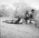 Two men lying on the ground, with two expedition members sitting on chairs next to them