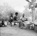 Two men lying on the ground, with three expedition members sitting next to them, a table and chest with food on it in front of them, belongings hanging in a tree near them