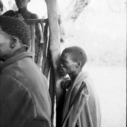 "/Gao Medicine" wearing a blanket over his shoulders, standing, leaning against a fence, with another man standing next to him