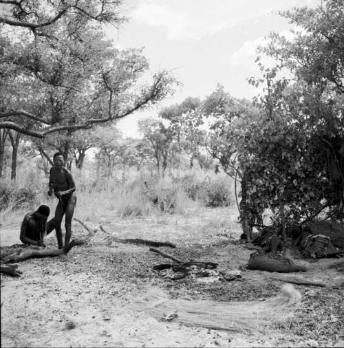 Two men working on arrows, one standing, one sitting, with a grass mat on the ground near them