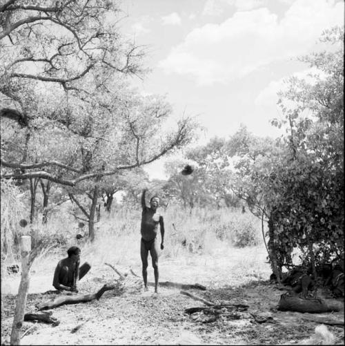 Man sitting, working on arrows, with another man standing next to him, gesturing