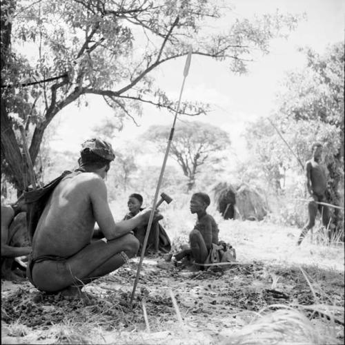 "/Gao Music" squatting with another man, holding their hunting equipment, including a small axe, with an assegai stuck into the ground next to them, two women sitting behind them