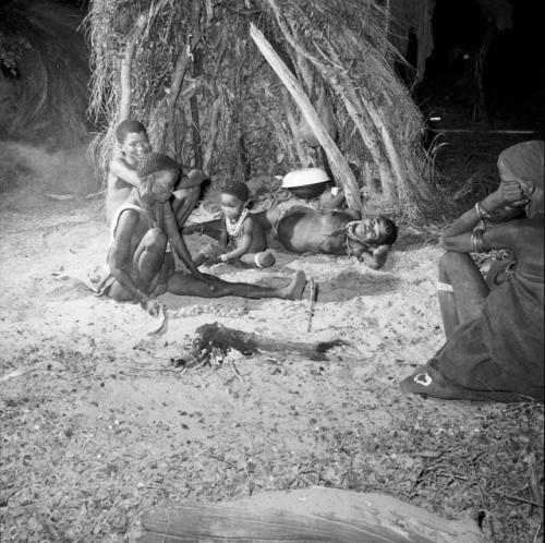 Woman stirring nuts in a fire with a fire paddle, sitting with her family in front of a skerm, an iron pot and enamel basin inside the skerm in the background